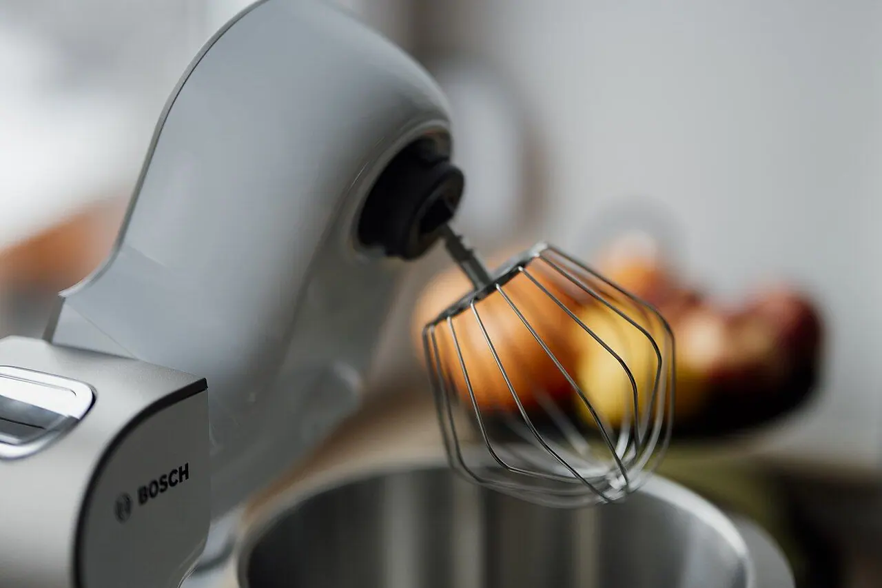 A kitchen mixer sits on a counter with a whisk attachment. In the background, fresh apples are placed in a bowl, indicating a baking task is about to start.